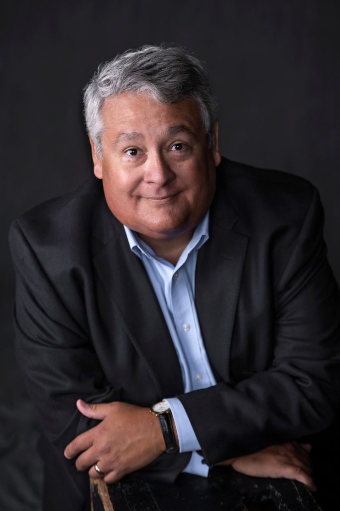 A middle-aged man with gray hair wearing a dark suit jacket and light blue shirt smiles softly while posing with his arms crossed against a dark background.