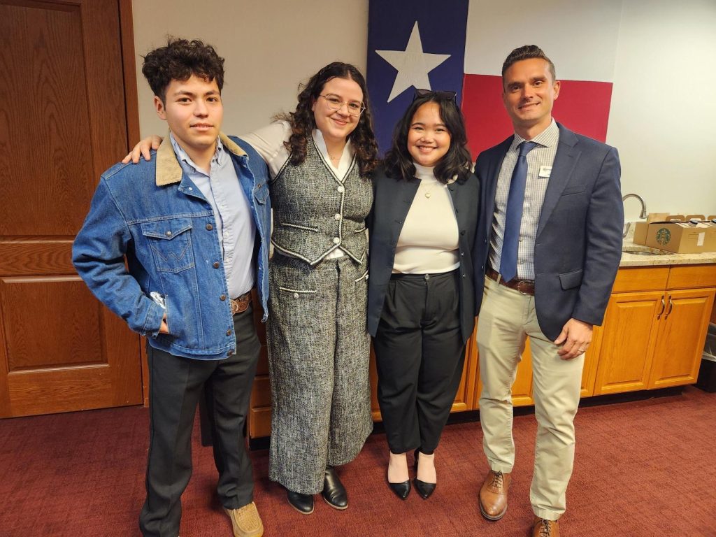 Four people stand side by side, smiling for a group photo in an office setting with a Texas flag on the wall behind them. They are dressed in business or business-casual attire.