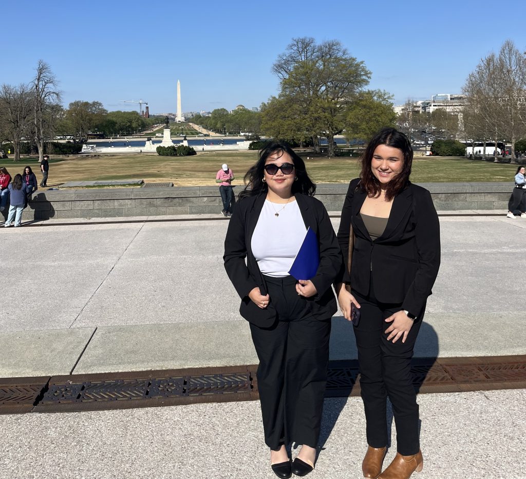 Two women in business attire stand outside on a sunny day with the Washington Monument visible in the background. Trees and people are scattered across the grassy area behind them.