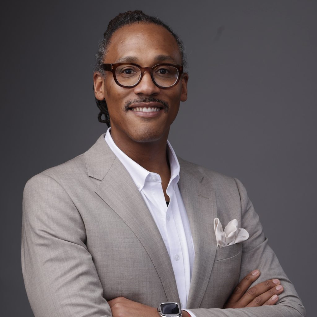 A man wearing glasses and a light-colored suit jacket with a pocket square stands against a gray background, smiling with arms crossed.