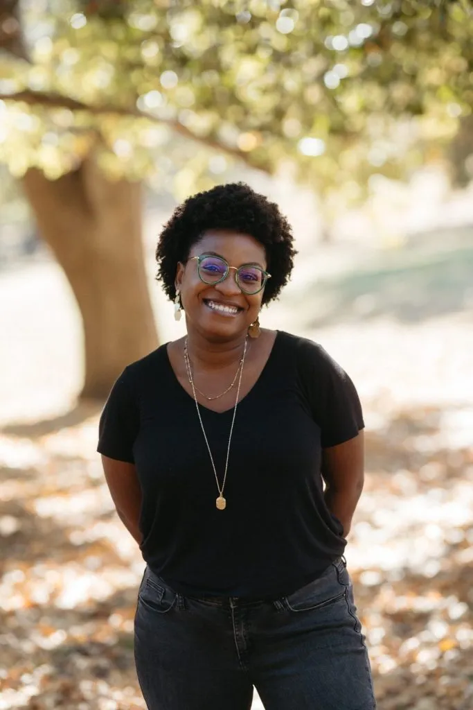 A woman with short curly hair, glasses, and gold jewelry smiles while standing outdoors in front of trees and fallen leaves, wearing a black t-shirt and jeans on a sunny day.