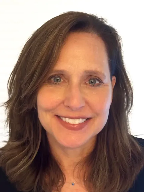 A woman with shoulder-length brown hair and blue eyes smiles at the camera against a light, blurred background. She is wearing a dark top and a delicate necklace.