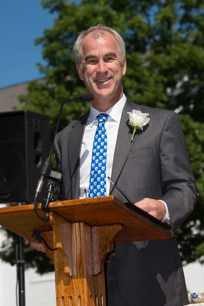 A smiling man in a suit and patterned blue tie stands at an outdoor podium with microphones, wearing a white rose boutonniere. Trees and a building are visible in the background.