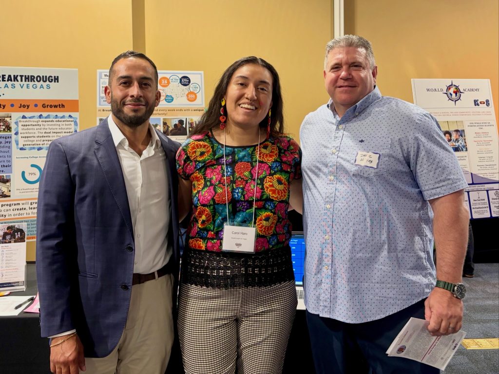 Three people stand smiling together at an indoor event, with educational posters displayed behind them. The person in the center wears a colorful embroidered shirt and name tag, while the others wear collared shirts and suit jackets.