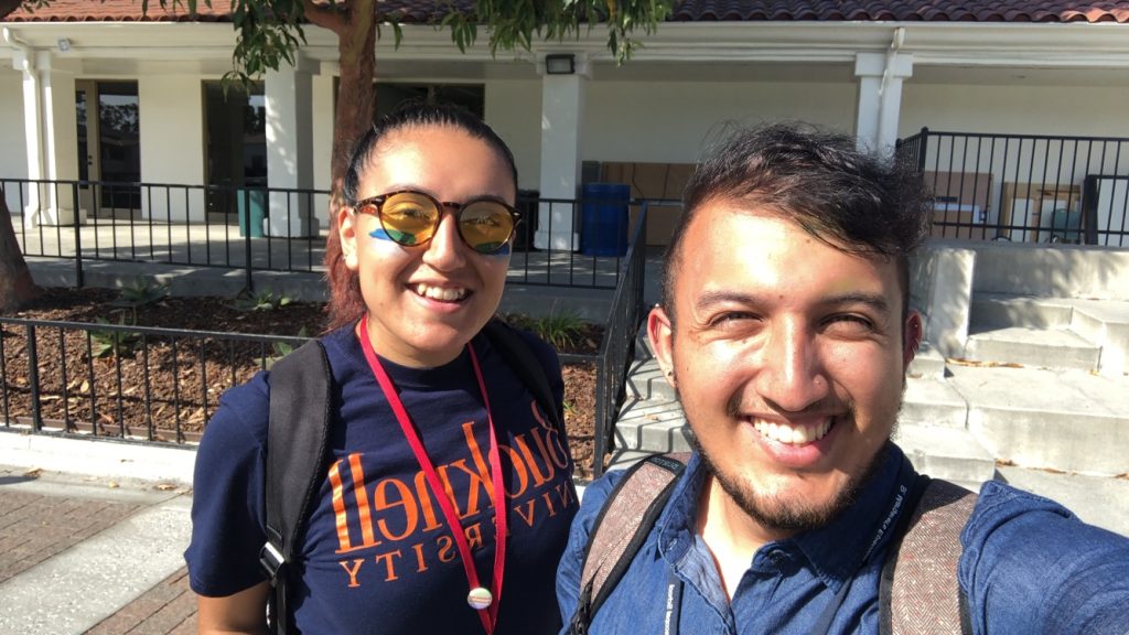 Two people smiling outdoors on a sunny day, both wearing backpacks. One person wears round sunglasses and a Bucknell University shirt, while the other wears a blue shirt. They stand in front of a building with steps and a tree.