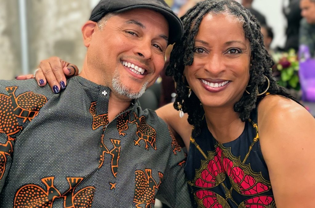 A smiling man and woman pose together, both wearing vibrant, patterned clothing with African-inspired designs. The man has a black cap and beard, and the woman has long, twisted hair. They look happy and close.