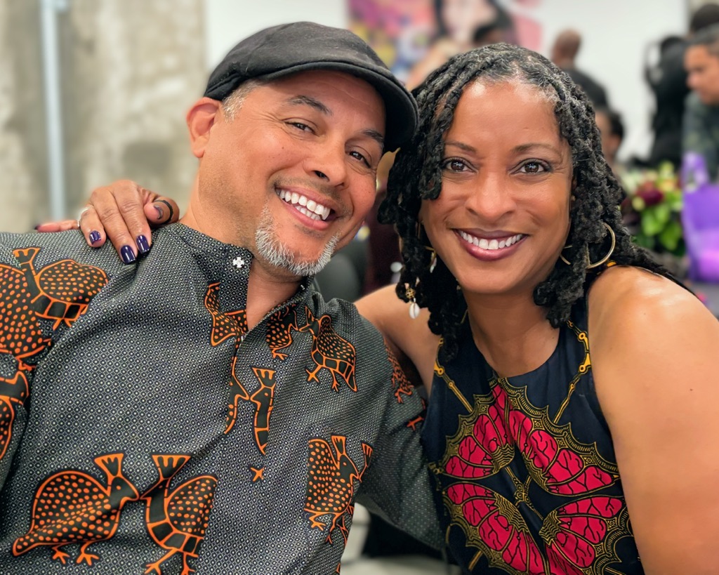A smiling man and woman pose together, both wearing vibrant, patterned clothing with African-inspired designs. The man has a black cap and beard, and the woman has long, twisted hair. They look happy and close.