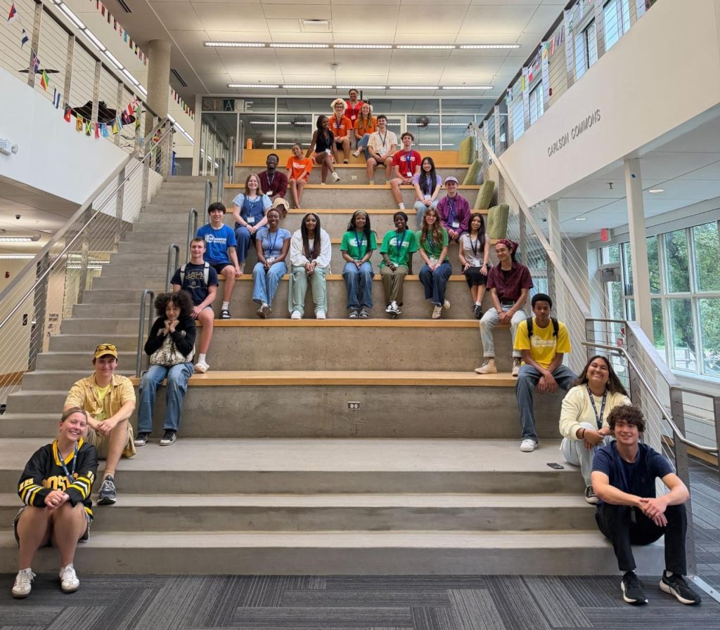 A diverse group of students wearing colorful shirts sits in rows on large indoor concrete steps in a modern school building with glass railings and banners above.