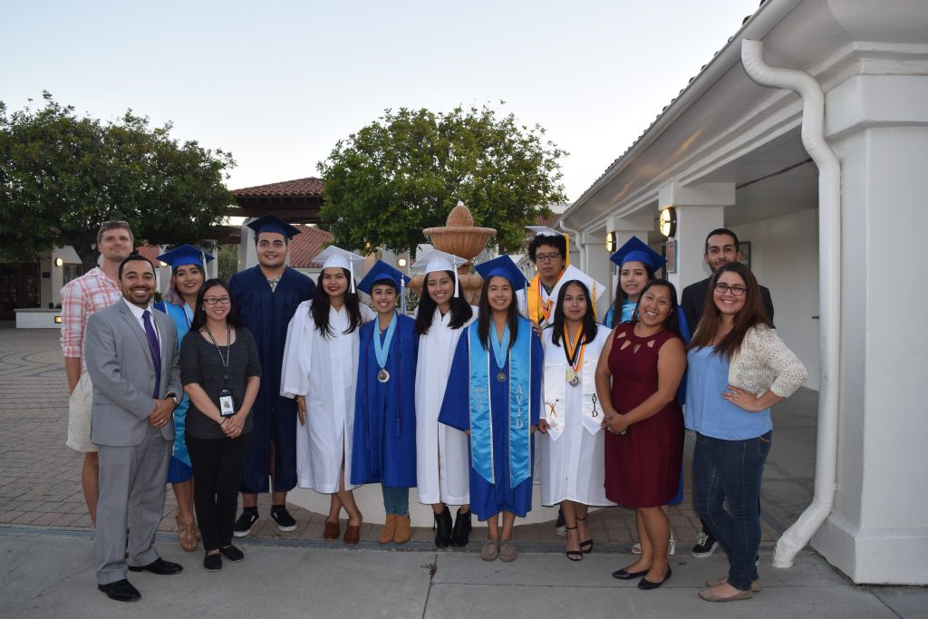A group of graduates in caps and gowns pose outdoors with smiling teachers and staff. The setting is a courtyard with trees and a white building in the background.