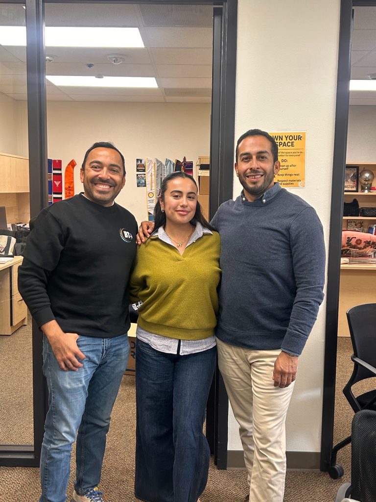 Three people stand indoors, smiling at the camera. Two men are on either side of a woman. Office furniture, boxes, bookshelves, and posters are visible in the background.