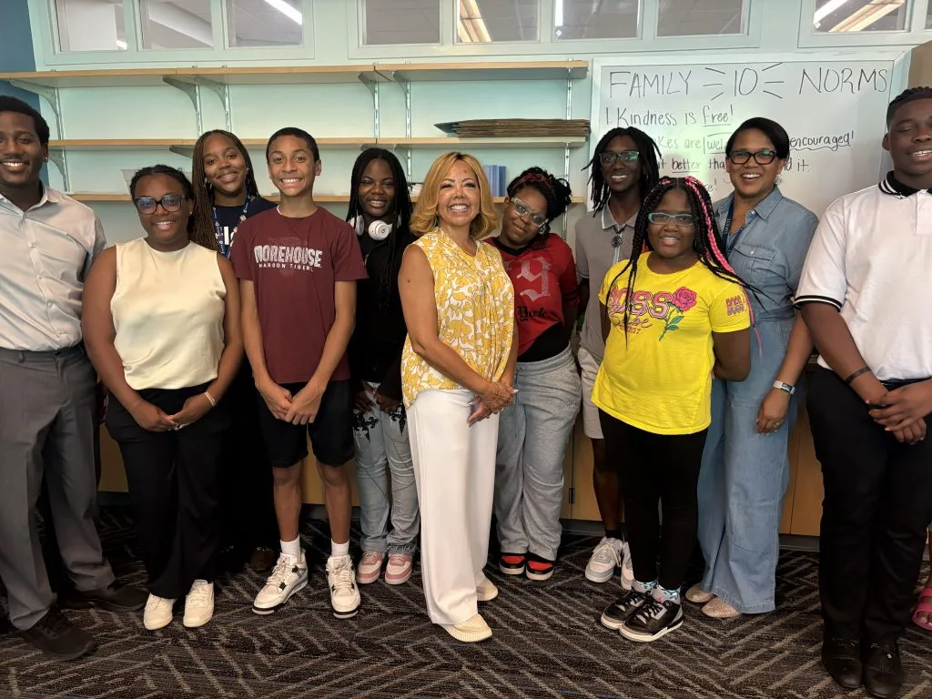 A group of students and adults stand together smiling in a classroom. Behind them is a whiteboard with handwritten notes and the words FAMILY = 1O NORMS at the top.
