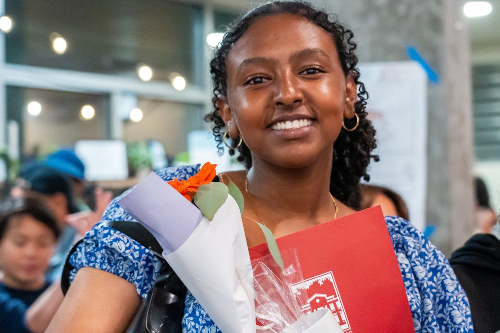A young woman in a blue patterned dress smiles, holding a bouquet of flowers and a red folder. She stands indoors, with blurred people and windows in the background.