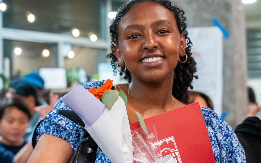 A young woman in a blue patterned dress smiles, holding a bouquet of flowers and a red folder. She stands indoors, with blurred people and windows in the background.