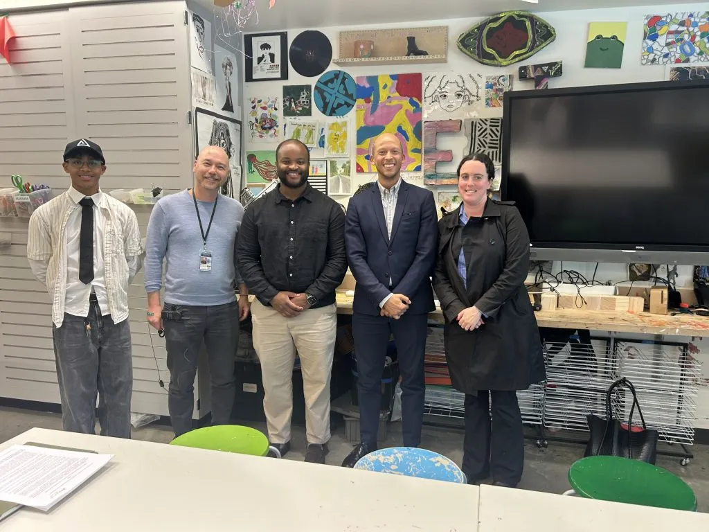 Five adults pose for a photo in an art classroom with colorful student artwork on the wall behind them, and art supplies on desks nearby in a school setting.