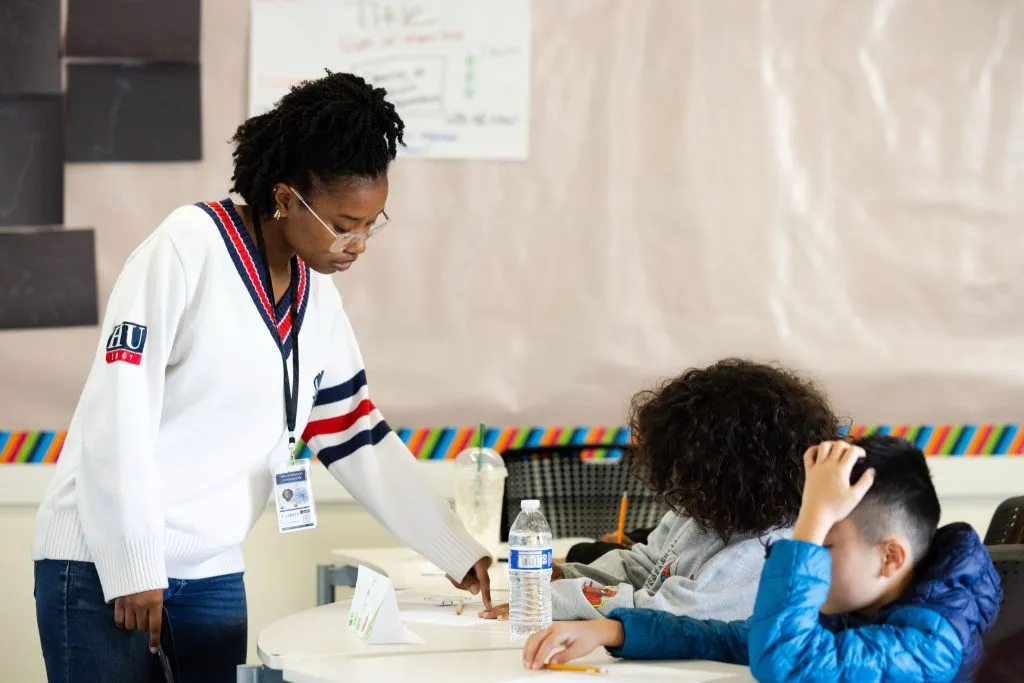 Teacher in a white sweater hands papers to students at a classroom table, guiding a hands-on activity.