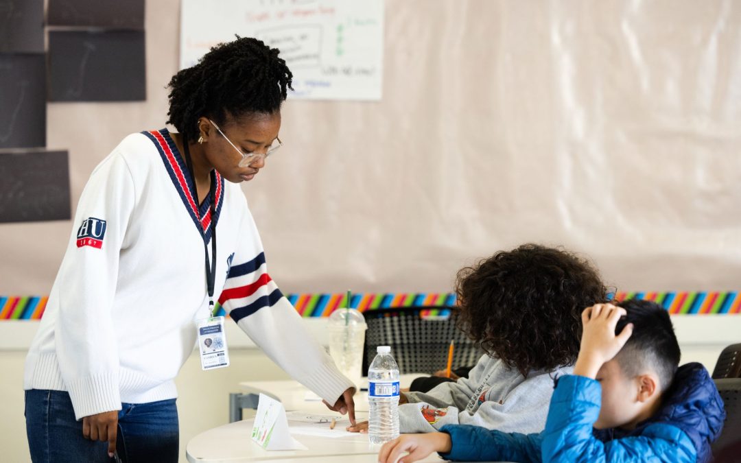 Teacher in a white sweater hands papers to students at a classroom table, guiding a hands-on activity.