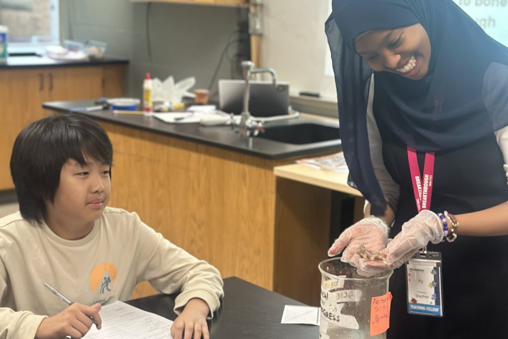 A smiling teacher wearing gloves shows a specimen to a student, who is seated at a desk and writing on a worksheet in a science classroom.
