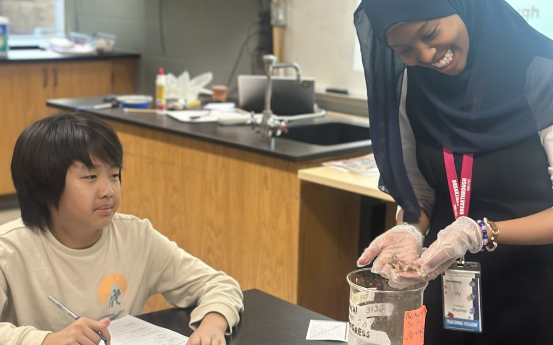 A smiling teacher wearing gloves shows a specimen to a student, who is seated at a desk and writing on a worksheet in a science classroom.