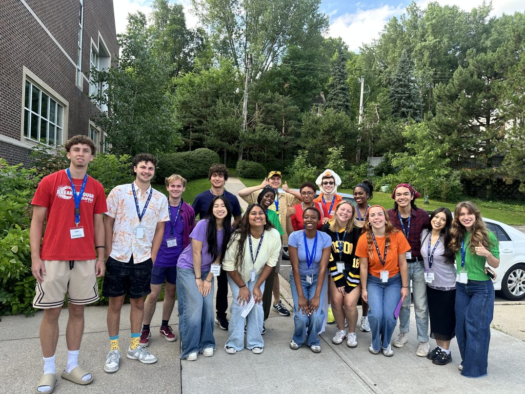 A group of smiling young adults pose together outdoors near a building, wearing colorful casual clothes and lanyards, with trees and greenery in the background.