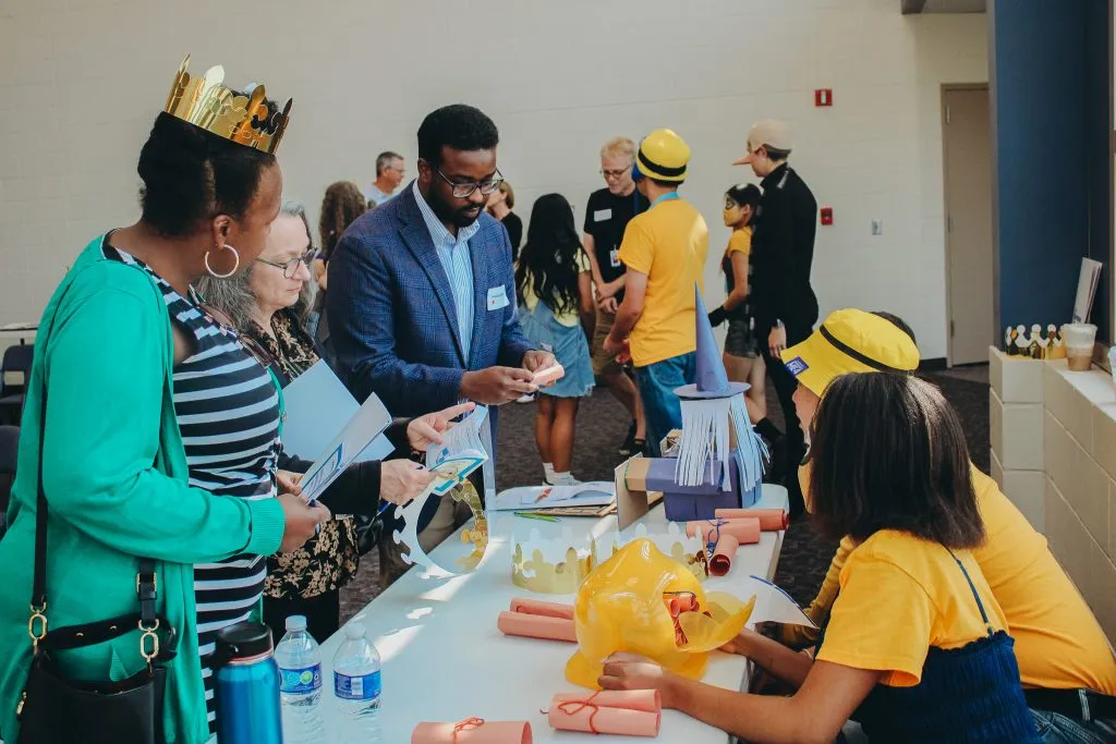 People line a long table filled with paper crafts as they look through guides; a man in a blue blazer stands center helping attendees.