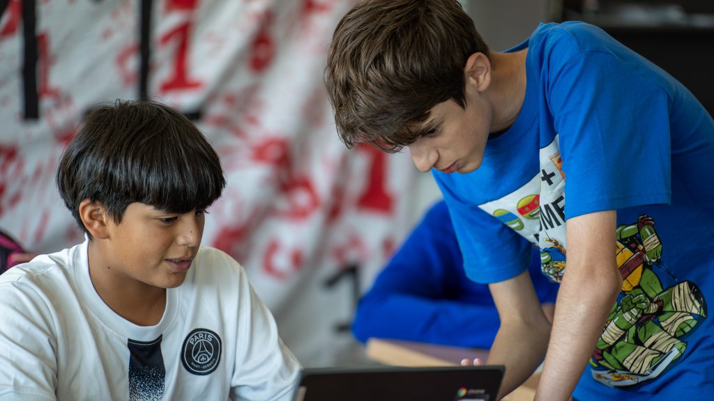 Two boys work together at a table, one seated and one standing, looking at a laptop screen. The background is blurred with red and white colors.